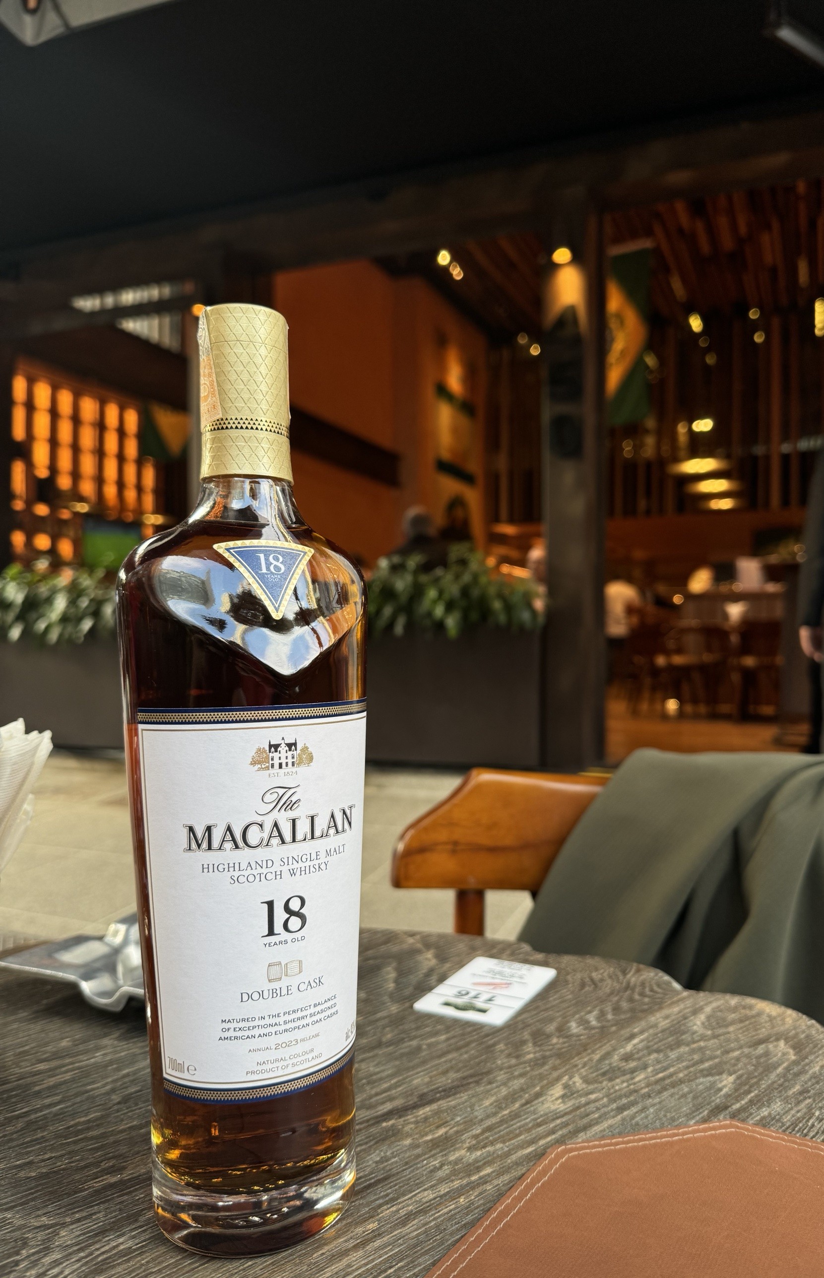 Garrafa de The Macallan 18 anos Double Cask numa mesa de madeira, com a fachada de ripado de um restaurante ao fundo