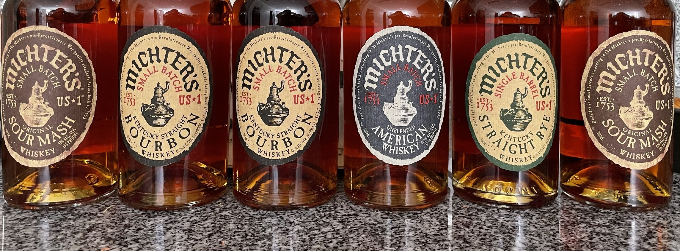 Seis garrafas de Michter&rsquo;s US★1 alinhadas numa bancada de granito, com os rótulos à vista