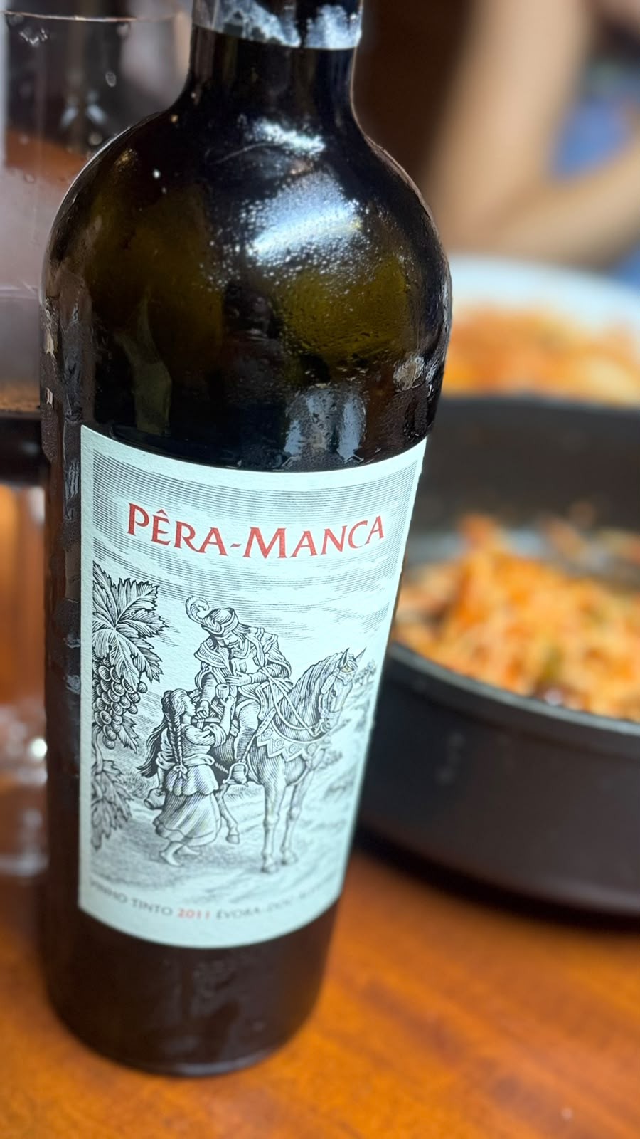 Garrafa de Pêra-Manca tinto 2011 sobre uma mesa de madeira, com um tacho de comida ao fundo