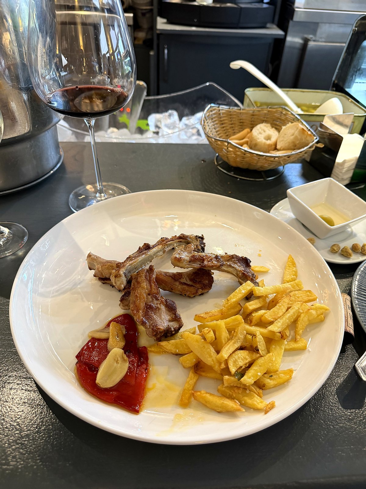 Costeletas de cordeiro com pimento vermelho assado, alho e batatas fritas, com uma taça de vinho tinto e um cesto de pão ao fundo