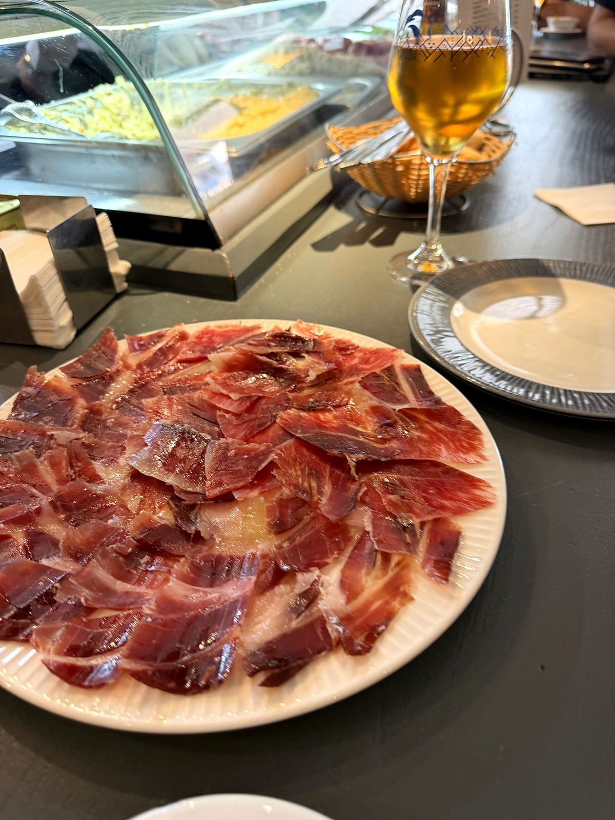 Prato de presunto ibérico fatiado num balcão, com uma taça de cerveja e um cesto de pão ao fundo