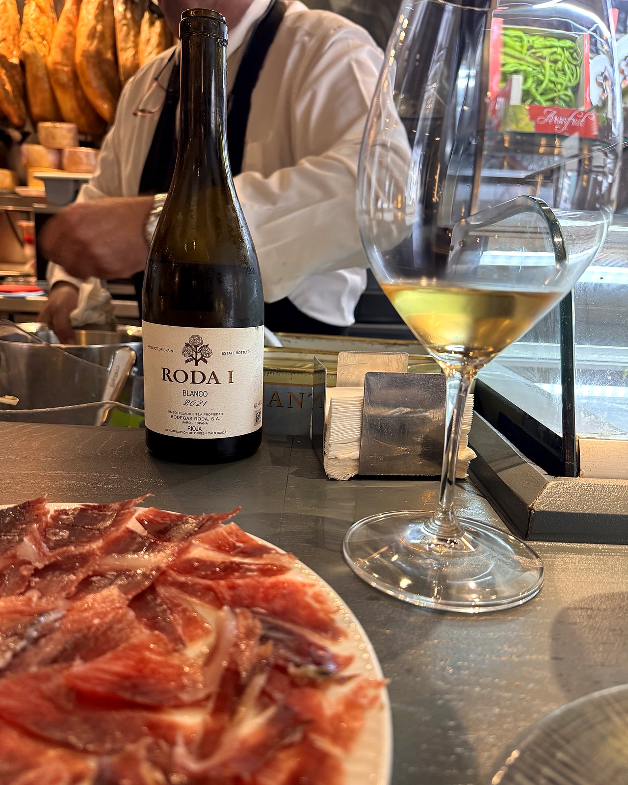 Garrafa de Roda I Blanco 2021 e uma taça de vinho branco em um balcão, ao lado de um prato de presunto ibérico