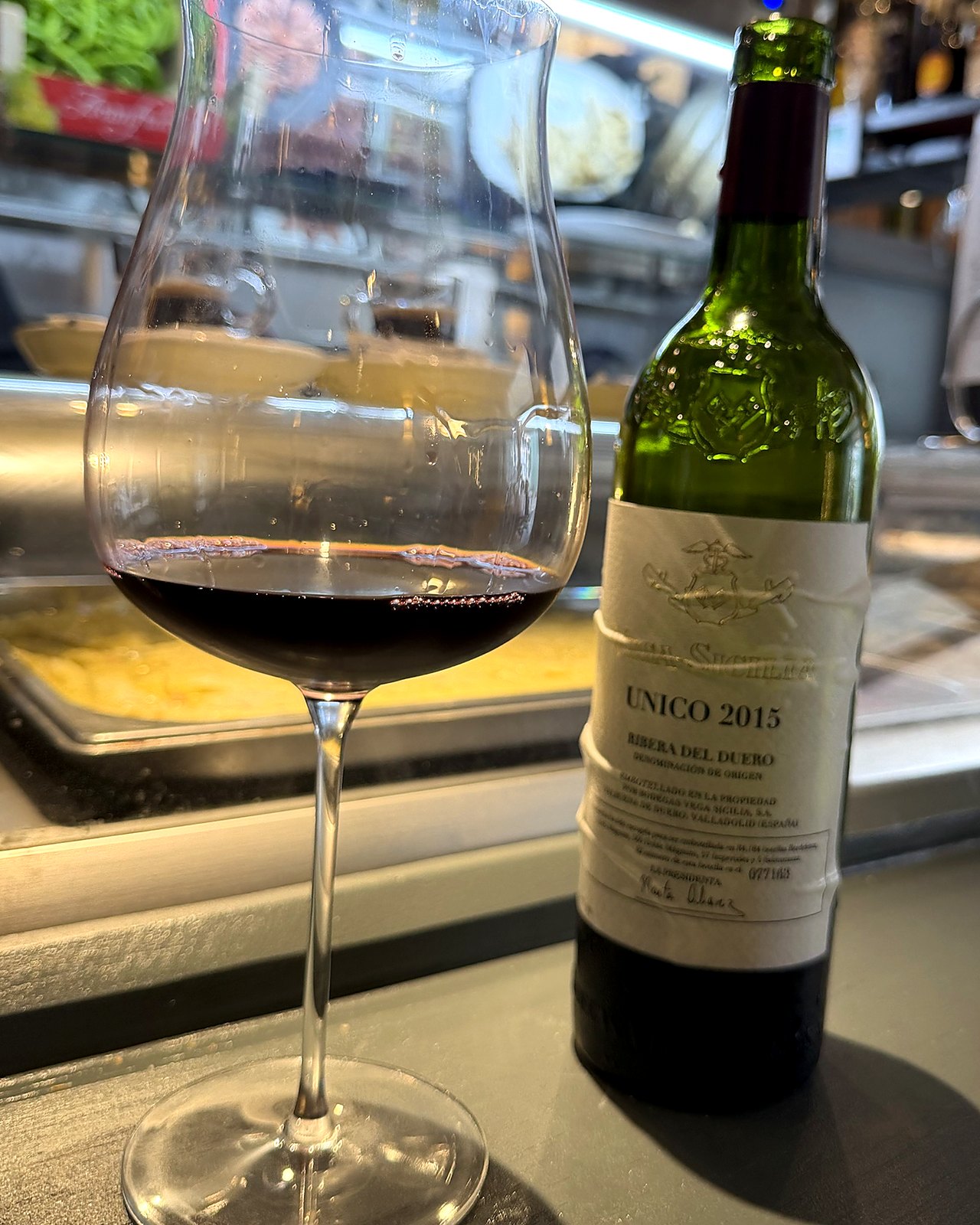 Taça de vinho tinto ao lado de uma garrafa de Vega Sicilia Único 2015 num balcão de bar